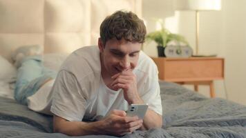 Caucasian man lying on bed using mobile phone smartphone cellphone texting smiling guy male communication online web technology internet social media networks bedroom home enjoy sms chatting gadget video