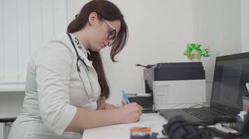 Physician in general practice completing medical card. General practitioner in a white coat does paperwork at the workplace with a laptop, writes notes, prepares documents, reports, prescriptions. video