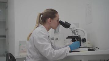 Female scientist using microscope while working in laboratory. Lab for modern medical research. Woman laboratory assistant looking in microscope analyzing samples biochemical substances, blood tests. video