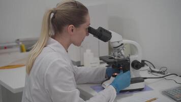 Female scientist using microscope while working in laboratory. Lab for modern medical research. Woman laboratory assistant looking in microscope analyzing samples biochemical substances, blood tests. video