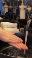 hand touches the water and washes hands with soap. A black sink with a silver faucet next to an oval mirror and a shelf with hand paper towels. Close-up video