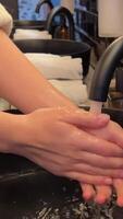 Washing hands in a black sink. black toilet is in a bathroom with a black tile wall. hand touches the water and washes sink next to an oval mirror and a shelf with hand paper towels. Close-up video
