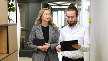 homem caminhando dentro uma corredor enquanto segurando uma prancheta quando a colega de escritório cumprimentar ele. dois escritório trabalhadores andar juntos dentro uma corredor, mulher com ipad partilha Ideias sobre dele Arquivo dentro prancheta. video