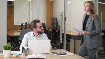A serious businessman working on his laptop. Officemate asking the man sitting on his chair about handheld printed marketing data reports. video