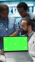 Vertical Team of doctors consulting analytics next to green screen on laptop in medical cabinet, examining x ray scan results to find the diagnosis. Licensed medics review hospital records at desk. Camera B. video