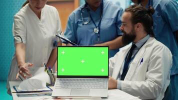 Team of doctors consulting analytics next to green screen on laptop in medical cabinet, examining x ray scan results to find the diagnosis. Licensed medics review hospital records at desk. Camera A. video