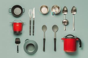 Colorful kitchen utensils arranged creatively on a teal surface, featuring red pots and diverse metal tools photo