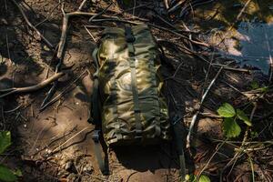 A rugged green backpack resting on sandy terrain near a tranquil stream during a sunny afternoon photo