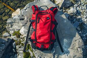 A vibrant red backpack rests on rugged stones by the water's edge under the bright afternoon sun photo
