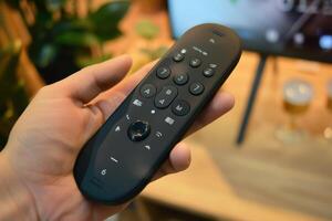 Hand holding a remote control in a cozy indoor setting with plants and a television in the background photo