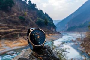 A close-up of a sleek watch resting on a rock by a tranquil river surrounded by mountains during the day photo