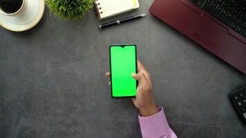 A man is using a smartphone with a green screen on a desk with a laptop and a cup video