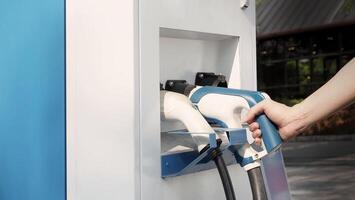Hand holding EV charging plug in front of camera with blurred background of outdoor. Driver of electric car hold batteries charger plug. Sustainable energy powered electric charging station background photo