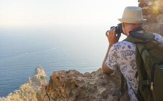 A young man with a backpack takes pictures of the landscape on top of a mountain. Back view. photo