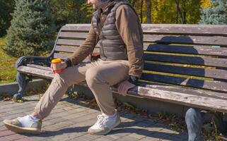 A young man sits on a bench in an autumn park, drinks coffee, rests, relaxes. Urban lifestyle concept. photo