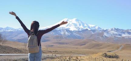 A happy female traveler with a backpack behind her back and her arms raised up admires the snow-covered Elbrus on a clear autumn day. View of Elbrus, North Caucasus, Russia photo