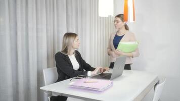 A woman at a desk using a laptop while another woman stands behind her video