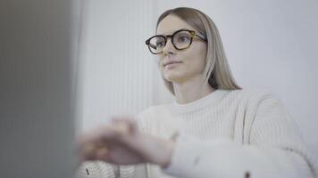 A happy woman with glasses sits at a desk, smiling while working on a laptop video