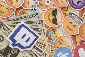 KYIV, UKRAINE - APRIL 20, 2024 Twitch logo and many usd money bills. Symbol of famous social network platform and dollar currency banknotes on lot of emoji reactions photo