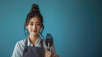 Portrait of a Smiling Young Woman in Apron Holding a Spatula Against Blue Background photo