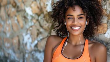 Smiling Fitness Woman in Bright Orange Top with Earbuds Leaning Against a Stone Wall photo