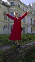 uma alegre mulher dentro uma brilhante vermelho vestir poses com braços elevado dentro frente do uma grande, abandonado mansão. a tiro captura dela expressivo posição a partir de uma baixo ângulo, enfatizando a contraste entre dela video