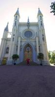 A woman in a flowing red dress runs towards the grand entrance of a Gothic church, captured from a low-angle perspective. The image emphasizes the contrast between her vibrant attire and the intricate video
