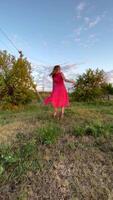 A joyful woman wearing a flowing pink dress spins around in a grassy field during sunset. The image captures her movement and happiness against a rural backdrop with trees and power lines. The warm video