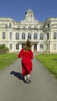 A woman in a vibrant red dress runs towards a grand, abandoned mansion under a bright blue sky. The image captures her from behind, highlighting the dynamic movement against the backdrop of the video