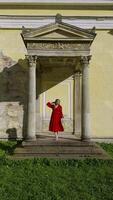 A woman in a red dress stands on the steps of a historic building, framed by classical columns and ornate architecture. The image captures her poised expression and the contrast between her vibrant video