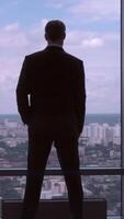 Full length back view of successful businessman in suit standing in office with hands on waist, CEO. Businessman from the back in front of a city view on the window video