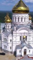 Huge white church. Drone view. Clip. A green landscape with a temple with golden domes standing in the middle and behind it a green forest and walking Christians and a blue sky with clouds. video