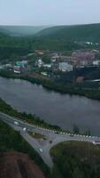Top view of small factory town by river. Clip. Beautiful landscape with highway and river near town with factory in wooded area. Factory town by river with bridge in hilly forest area in summer video