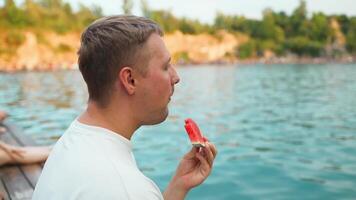 Handsome young man in stylish clothes sits enjoying a summer day at the lake and eating watermelon. video