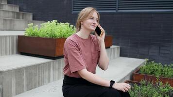 Caucasian 30s blonde woman in casual clothes siting near office building from outside, female smiling and using phone, freelancer typing message and browsing internet pages. video