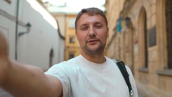 Handsome man taking selfie with smart mobile phone device or call by camera on city street. Happy tourist visiting Krakow old town. video