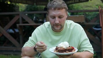 Handsome young man in stylish clothes sits and relaxes in a cafe. Male eats and enjoys a delicious chocolate cake with vanilla ice cream in a street cafe. video