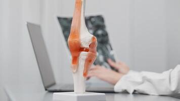 Orthopedic specialist analyzes imaging results in a modern office, using a laptop and knee joint model for detailed assessment. Showcases advanced orthopedic diagnostics and consultation video