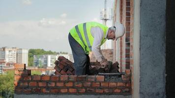 Mason Working on Brick Wall at Construction Site. A construction worker in a high-visibility vest is focused on building a brick wall on the upper levels of a construction site. video