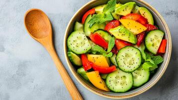 Overhead view of cucumber salad with bell pepper slices avocado chunks and chia seeds in a ceramic bowl with a wooden spoon beside it photo