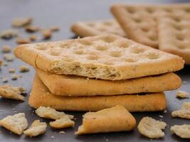 stack of square crackers with pieces and crumbs on slate gray background. Dry salt cracker cookies with fiber and dry spices photo