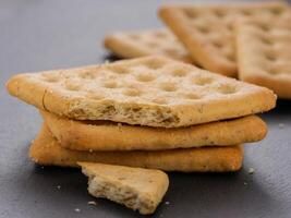 stack of square crackers with pieces and crumbs on slate gray background. Dry salt cracker cookies with fiber and dry spices photo