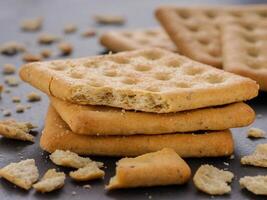 stack of square crackers with pieces and crumbs on slate gray background. Dry salt cracker cookies with fiber and dry spices photo