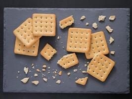 Square crackers with pieces and crumbs on slate gray background. Dry salt cracker cookies with fiber and dry spices. Top view or flat lay. photo