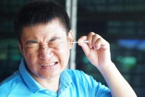 Asian man make face expression during using cotton bud stick to remove earwax. Concept, Health care. Use cotton swab in ear can hurt ear drums and hearing loss. Ear cleaning photo