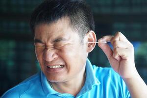 Asian man make face expression during using cotton bud stick to remove earwax. Concept, Health care. Use cotton swab in ear can hurt ear drums and hearing loss. Ear cleaning photo
