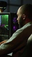 Vertical Smiling man playing games on gaming PC at computer desk, enjoying day off from work. Upbeat gamer battling enemies in online multiplayer shooter from neon lights ornate home, camera A video