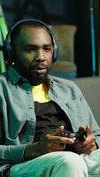vertical alegre homem vestindo fones de ouvido, a comemorar depois de vitória dentro videogame, néon aceso apartamento. pessoa em sofá lança punhos dentro ar a partir de excitação depois de ganhando jogos em console com controlador, Câmera uma video