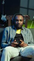 vertical alegre homem vestindo fones de ouvido, a comemorar depois de vitória dentro videogame, néon aceso apartamento. pessoa em sofá lança punhos dentro ar a partir de excitação depois de ganhando jogos em console com controlador, Câmera b video