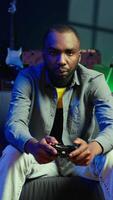 vertical jogador dentro rgb aceso casa jogos com controle de video game e desfrutando relaxamento tempo. homem sentado em sofá dentro néon iluminado vivo sala, jogando videogames em console usando controlador, Câmera b